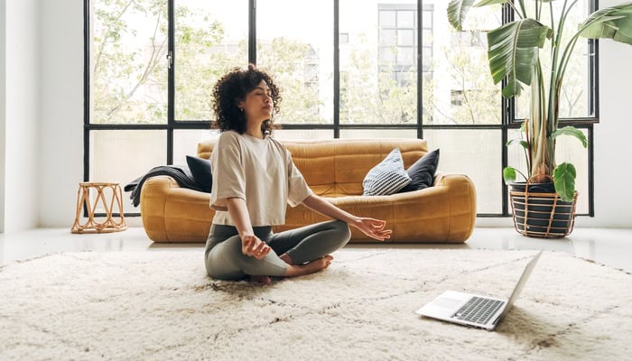 A Black woman sits on the floor in her bedroom in a yoga position, her eyes closed, in front of her laptop.
