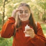 A woman wearing a light, red coat applies sunscreen from a tube to her face as she walks through the woods.