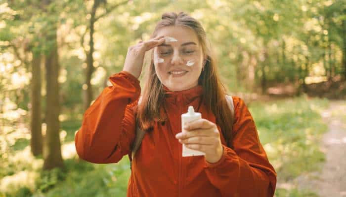 A woman wearing a light, red coat applies sunscreen from a tube to her face as she walks through the woods.