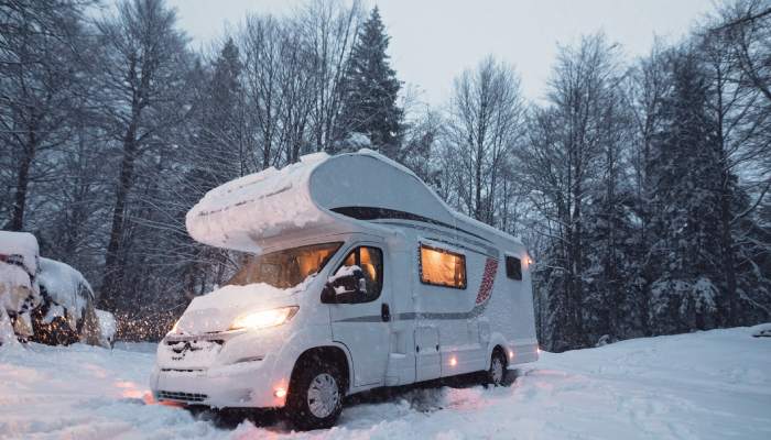 An RV sits at a snowy campsite surrounded by tall, leafless trees. The lights shine in the van and the falling snow glitters.