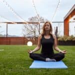 A woman wearing black athletic gear sits on a blue yoga mat with her legs crossed and eyes closed. String lights are above her.
