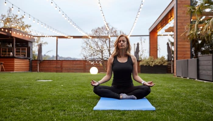 A woman wearing black athletic gear sits on a blue yoga mat with her legs crossed and eyes closed. String lights are above her.