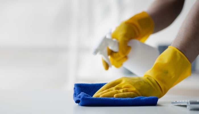 A close-up shows a person wearing yellow latex gloves spraying cleaner onto a white surface and wiping it with a blue rag.
