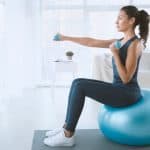 A woman in leggings and a tank top sitting on a blue exercise ball and holding dumbbells with a couch in the background.