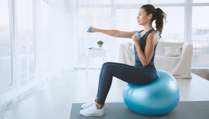 A woman in leggings and a tank top sitting on a blue exercise ball and holding dumbbells with a couch in the background.