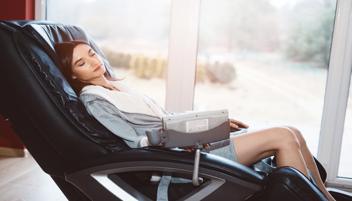 A woman is resting in a black massage chair that sits next to a sliding glass door. She has her eyes closed.