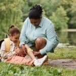 A mother kneeling down to place a bandage on the shin of her daughter. Both are sitting in a green park area.