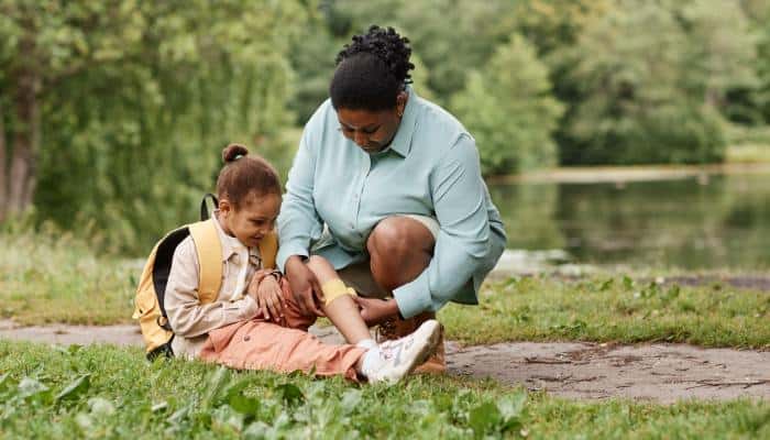 A mother kneeling down to place a bandage on the shin of her daughter. Both are sitting in a green park area.
