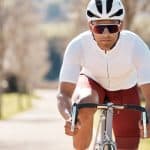 A man on a bicycle approaching the viewer in professional cycling gear, a white helmet, and large sunglasses.
