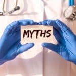 Close-up of a doctor's hands wearing blue examination gloves and holding a paper card with the word "Myths" on it.