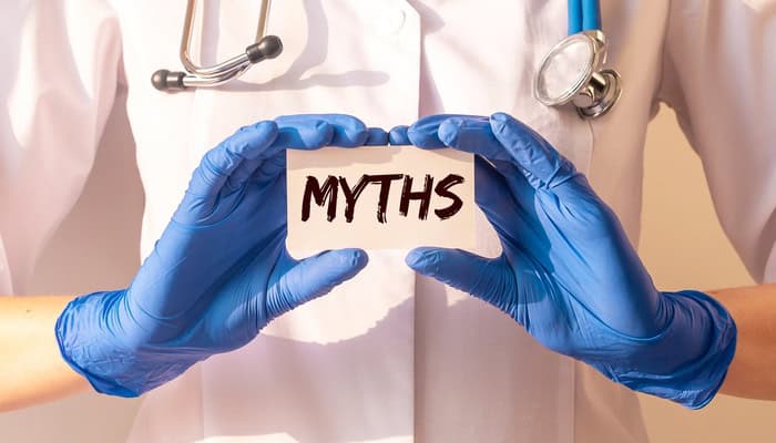 Close-up of a doctor's hands wearing blue examination gloves and holding a paper card with the word "Myths" on it.
