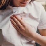 A young woman wearing a simple white t-shirt holding her hand to her chest, her face cast downward.