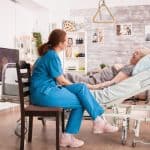 An older woman lays in a hospital bed set up in the middle of her living room. A nurse sits beside her in a chair.