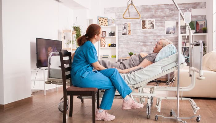 An older woman lays in a hospital bed set up in the middle of her living room. A nurse sits beside her in a chair.