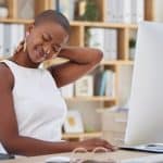 A woman in a sleeveless shirt sits in front of a desktop computer while rubbing the back of her neck in discomfort.