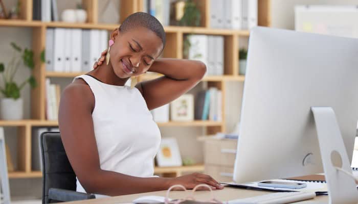 A woman in a sleeveless shirt sits in front of a desktop computer while rubbing the back of her neck in discomfort.