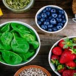 A bowl of spinach, blueberries, and strawberries sit on a wooden table next to different nuts and bananas.