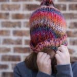 A woman pulling a colorful winter beanie over her face. She wears a gray sweater and stands in front of a brick wall.