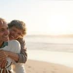 A senior couple standing on a clear beach. Behind the man, the woman has her arms wrapped around him.
