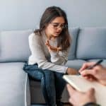 A young therapy patient sits on the couch across from the therapist as she looks down and talks with her hands.
