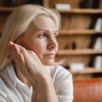 An aging woman sits on a leather sofa as she stares into the distance. She has a stressed look on her face.