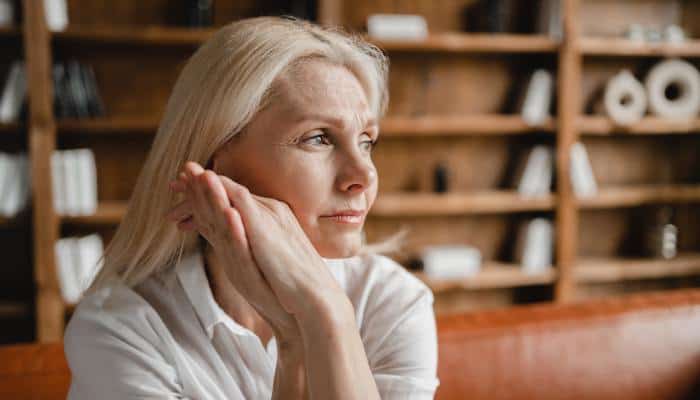 An aging woman sits on a leather sofa as she stares into the distance. She has a stressed look on her face.