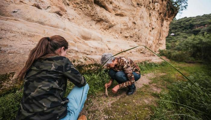 A mature woman and a young adult inspecting the ground of a trail while wearing camouflage and exploring outdoors.