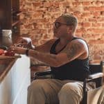 A person sits in their wheelchair next to a kitchen counter. They cut up vegetables on a cutting board.