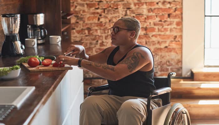 A person sits in their wheelchair next to a kitchen counter. They cut up vegetables on a cutting board.