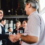 A young professional shaking hands with an older person in business casual as they both meet and greet at an event.