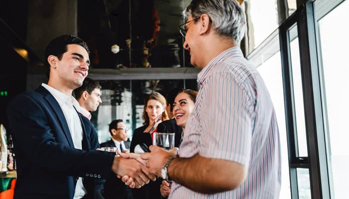 A young professional shaking hands with an older person in business casual as they both meet and greet at an event.