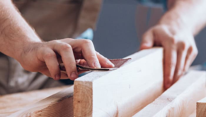 A woodworker's hands using a piece of sandpaper to sand down a long piece of wood in between two other pieces.