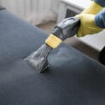 A close-up of a couch cushion being professionally cleaned by a cleaner in a suit. They wear yellow gloves to steam the couch.