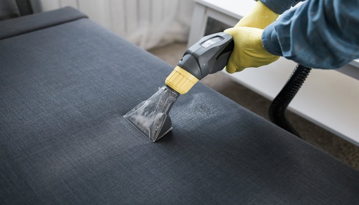 A close-up of a couch cushion being professionally cleaned by a cleaner in a suit. They wear yellow gloves to steam the couch.