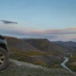 An off-roading car parked on a muddy cliff overlooking a river and mountains. A forest is also visible in the distance.