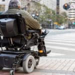 A person sitting in an electric wheelchair outside. The wheelchair is parked in front of a crosswalk on a busy street.
