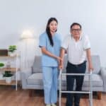An elderly man and his caregiver standing in front of a couch. The man is leaning his weight on the walker in his hands.