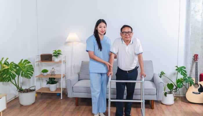 An elderly man and his caregiver standing in front of a couch. The man is leaning his weight on the walker in his hands.