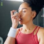 A woman wearing a workout shirt is closing her eyes and holding a white inhaler up to her mouth at the gym.