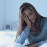 A woman sitting awake on a bed at night with her head in her hands in a dark bedroom illuminated by dim light.