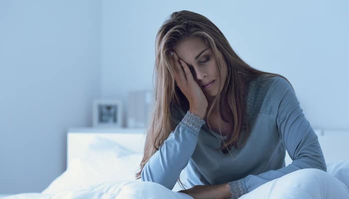 A woman sitting awake on a bed at night with her head in her hands in a dark bedroom illuminated by dim light.