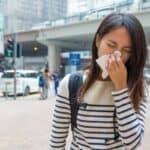 A young woman blows her nose with a tissue while standing in a plaza at a busy urban center on a cloudy day.
