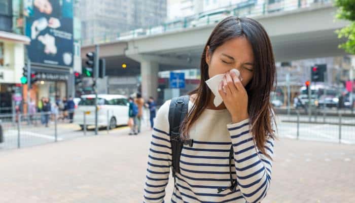 A young woman blows her nose with a tissue while standing in a plaza at a busy urban center on a cloudy day.